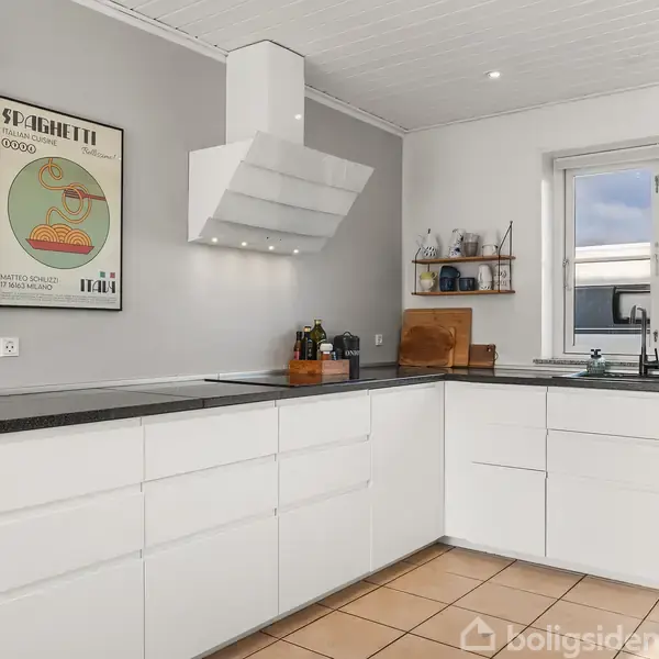 Hvidt køkken med sorte bordplader, vægmonteret vinreol og plakat med tekst "SPAGHETTI". Køkkenredskaber står på bordet ved siden af et vindue. Gulvet er dækket af lyse fliser.