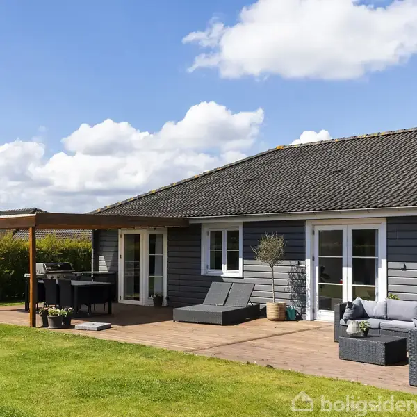 Hus med gråt træbeklædning står i en solrig have. En terrasse med havemøbler og en grill er placeret foran. Blå himmel og grøn plæne omgiver huset. Ingen tekst.