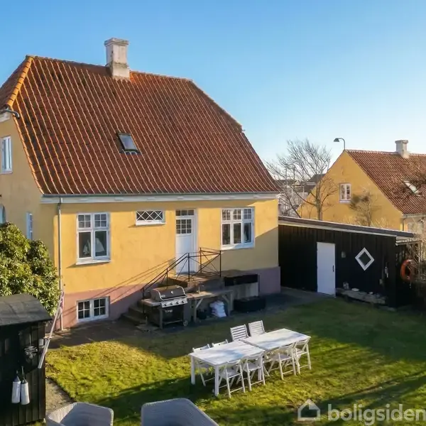 Gult hus med rødt tag står i en have med græsplæne. Der er et udendørs bord med stole, samt en gyngestativ ved siden af et hegn. Skyfrit, solrigt vejr.