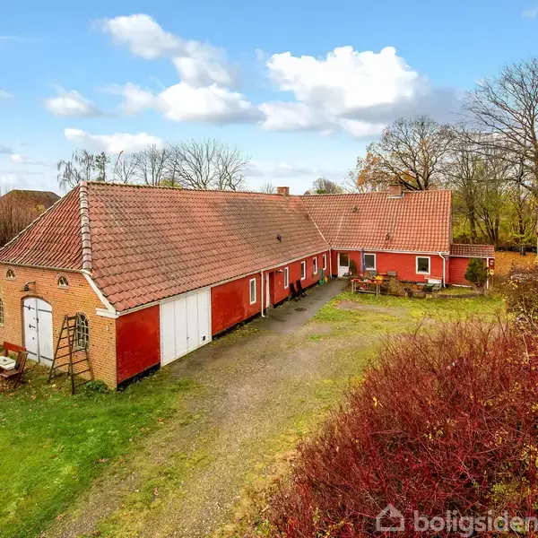 En stor, rødmalet landejendom med gavl og teglstenstag står stille. Grusvej foran fører til grønne områder med træer og buske omkring bygningen under en blå himmel med hvide skyer.