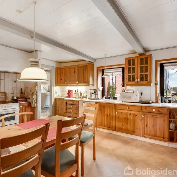 Et køkken med træmøbler og spisebord, omgivet af hvide vægge og et flisegulv. Vinduerne giver udsigt til en have. En lampe hænger over bordet.