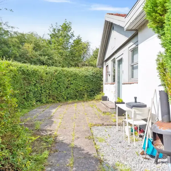 Haven har en terrasse med fliser og en havestol. En brændovn står ved siden af huset omgivet af grønne hække. Foran huset ligger smalle fliser langs en trimmed hæk.