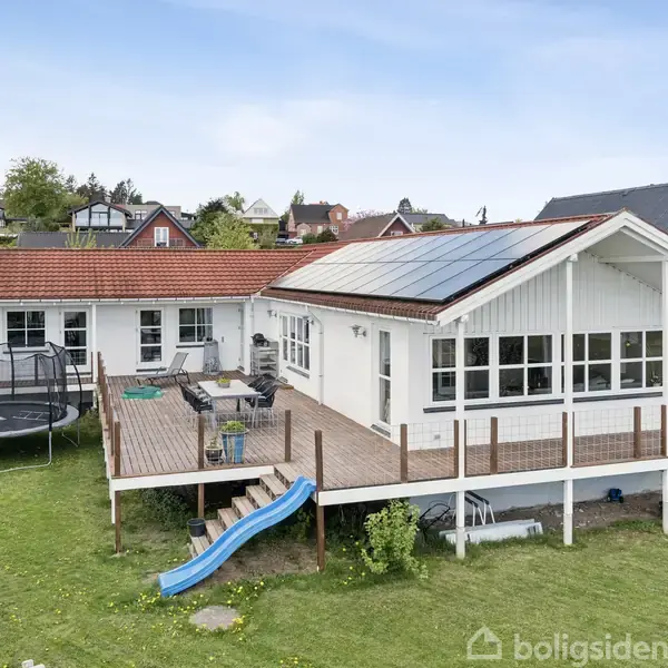 Hvidt hus med solpaneler på taget står på en stor grøn græsplæne. En træterrasse med møbler og en blå rutsjebane, med en trampolin til venstre.