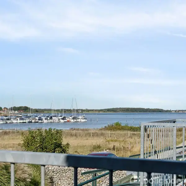 Både ligger stille ved en marina, omgivet af et grønt landskab, mens en blå himmel ses over en rolig vandflade.