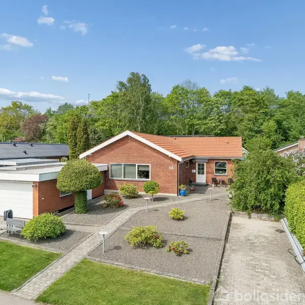 Rødt murstenshus med orange tegltag står stille omgivet af velplejet grusbelagt have og græsplæne. Garage ved siden af huset. Omgivet af grønne buske og træer under blå himmel.