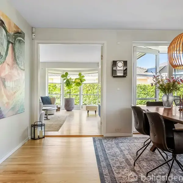 Et spisebord med sorte stole står under en trælampe i en stue. Ved siden af ses et vindue med udsigt til en terrasse. Et maleri hænger på væggen.