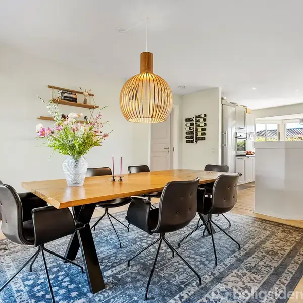 Et træbord står i en spisestue omgivet af seks sorte stole. Bordet har en vase med blomster. En stor lampe hænger over bordet. Stuen grænser op til et køkken.