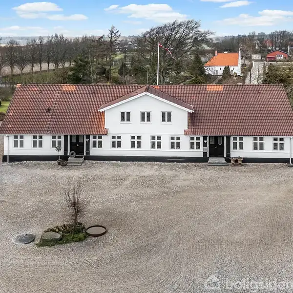 En hvid landvilla med rødt tag står i en grusbelagt gårdsplads. Omkringliggende bygninger ses, omgivet af marker og træer i baggrunden. Boligsiden-vandmærket ignoreres.