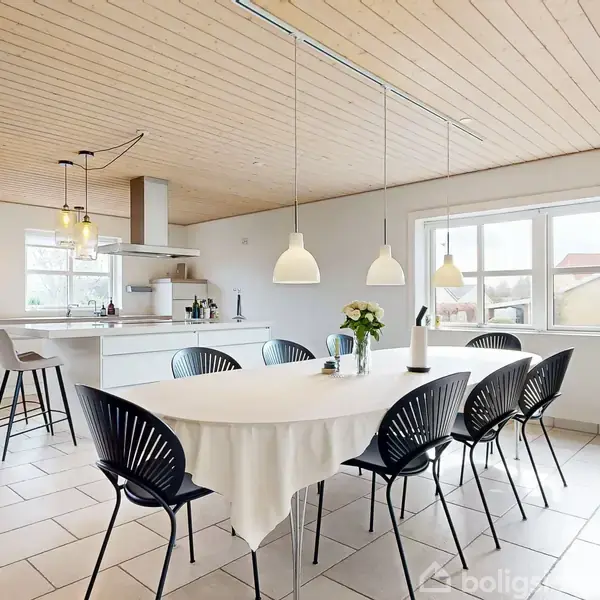En spisestue med et langt bord dækket med en hvid dug og sorte stole omkring. Køkkenet ses i baggrunden med høje taburetter. Store vinduer giver naturligt lys.
