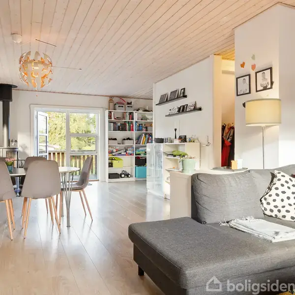 Stue med grå hjørnesofa, sofabord og gulvlampe; spisebord med stole står til venstre. Rummet har trægulv, hvide vægge, og lysindfald fra vindue og altandør.