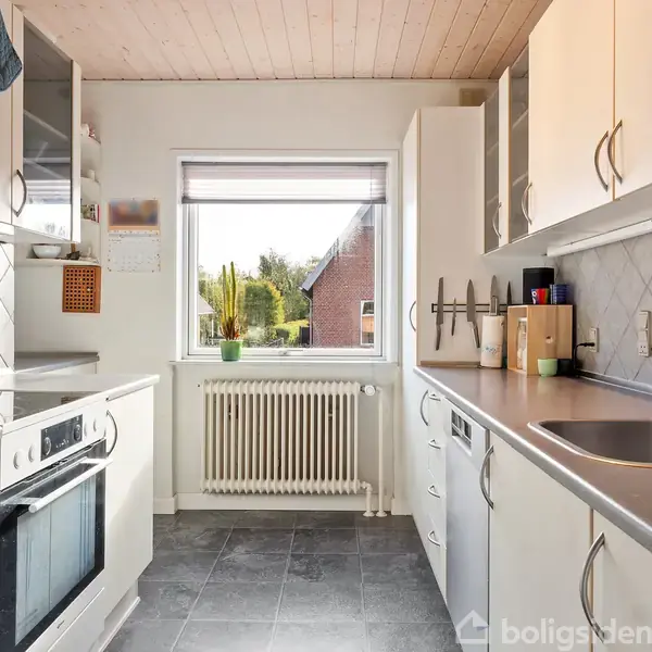 Et lyst køkken med hvide skabe og grå flisegulv, udstyret med en ovn og vask. Et vindue viser udsigt til en have med en kaktus som plante på vindueskarmen.