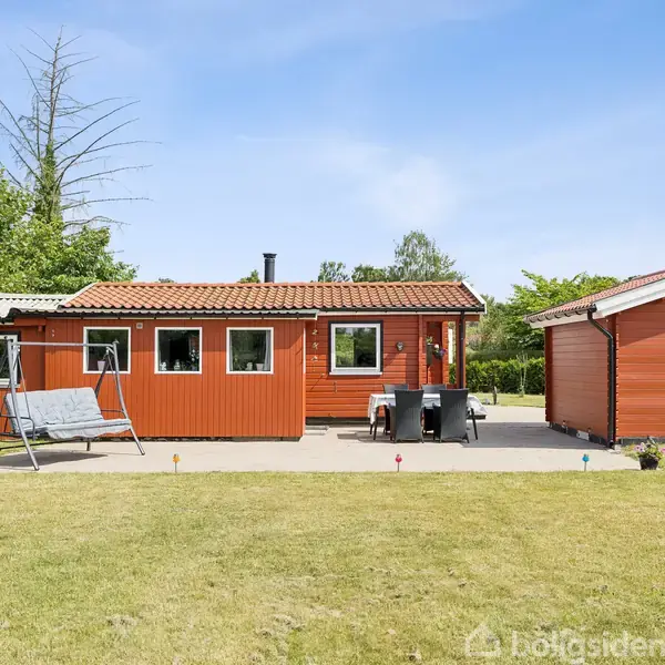 Et rødt sommerhus med hvide vinduer står stille på en terrasse. Ved siden af huset er et fritstående skur. Græsplæne og træer omgiver området under en klar blå himmel.