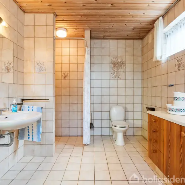 Et badeværelse med håndvask til venstre, toilet i midten og træskab med vask til højre. Flisebelægning på væg og gulv, bruser bag forhæng.