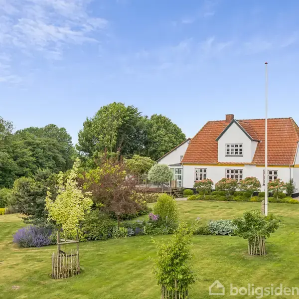 Et hvidt hus med rødt tag står i en frodig have. Træer og buske omkranser huset, som også har en terrasse. Flagstang i forgrunden.