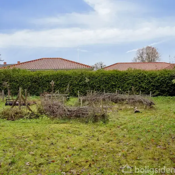 Grøn have, indeholdende enkle trækonstruktioner og nyligt klippet græsplæne, omgivet af høje, tætte buske. Villa med rødt tegltag i baggrunden under en klar blå himmel.