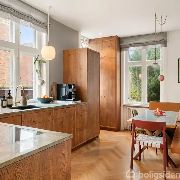 Køkken med træskabe, hvor en kaffemaskine står på en bordplade. Et spisebord med farverige stole og en bænk ved et vindue med udsigt til grønne omgivelser.