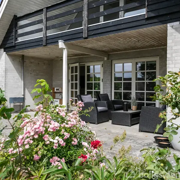 En terrasse med flettede havemøbler står foran et lyst hus med store vinduer. Blomstrende roser og grønne planter omgiver området, der er dækket af fliser.