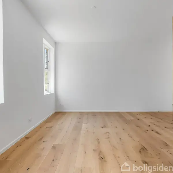 Tomt værelse med trægulv, stor garderobe til højre og to vinduer til venstre, udsigt til grønne træer udenfor. Hvidmalede vægge og minimalistisk indretning.