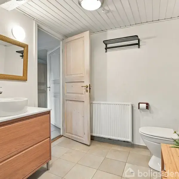 Et badeværelse med en vask på et træskab. En åben dør fører til en korridor. Der er et toilet ved siden af radiatoren, og et stort spejl hænger over vasken.