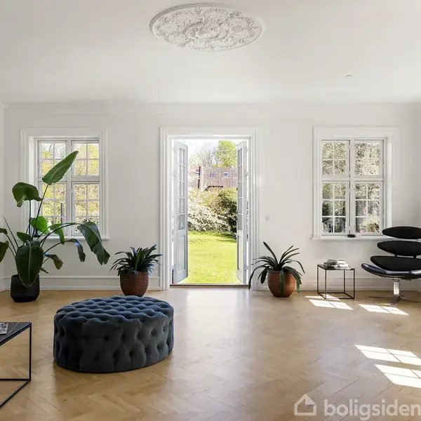 Stue med en mørkeblå puf i midten, omgivet af grønne planter. Dobbelt glasdør fører til en have med græs. Lyse vægge, parketgulv og moderne møbler.