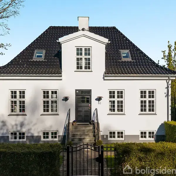 Hvid villa med sort tag, placeret bag en lille hæk og en indgangsport. Trapper fører til hoveddøren, omgivet af vinduer. Huset står i en grøn have med træer omkring.