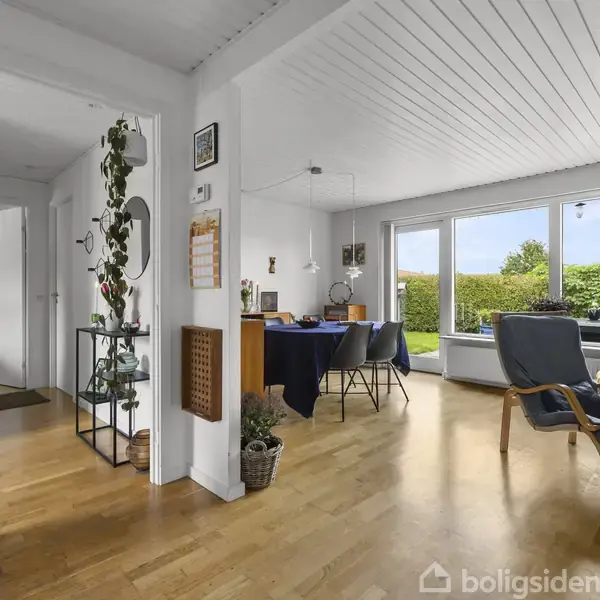 Stue med trægulv har en spiseplads til venstre med mørkeblå dug på bordet. Store vinduer til højre viser en velplejet have udenfor. Væggen har kalender og dekorationer.