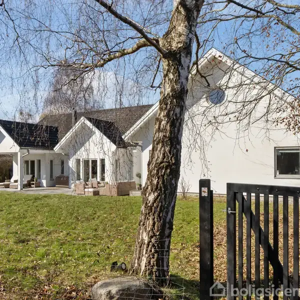 En hvid villa med sort tag står roligt i en grøn have. Foran huset er en terrasse med siddepladser, mens træer omgiver ejendommen. En sort låge markerer indgangen.