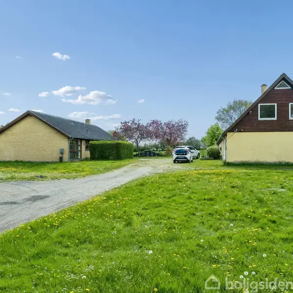 To gule huse står på en grøn græsplæne med en grusvej imellem dem. I baggrunden ses en bil parkeret nær nogle blomstrende træer under en blå himmel med skyer.