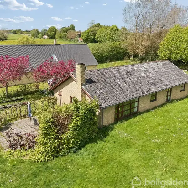 Et enkelt hus med skråtag ligger i en grøn have omgivet af træer og marker. En traktor ses til højre, og et større hus står i baggrunden ved blomstrende træer.