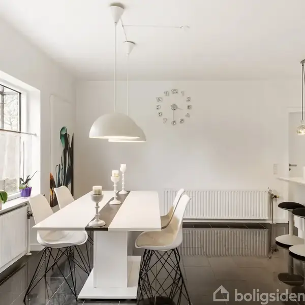 Et moderne, lyst køkken-alrum med et hvidt spisebord og seks stole. Bordet er dekoreret med lysestager. Store vinduer til venstre giver lys. En væg-ur hænger centralt.