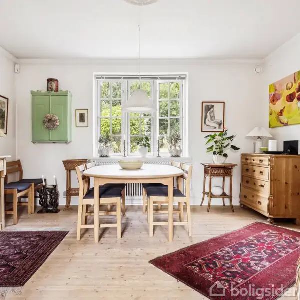 Spisebord med stole står i midten af et lyst rum. Omgivet af tæpper, skabe og et vindue med udsigt til grønne træer. Kunst og dekorationer pryder væggene.