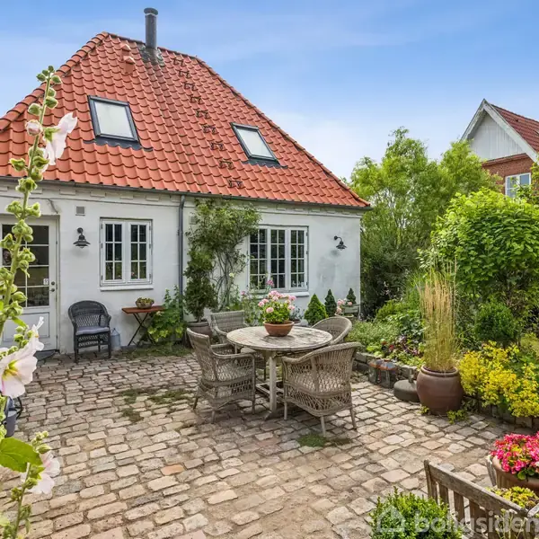 Hvidt hus med rødt tegltag står i en brostensbelagt gårdhave. Havemøbler og blomsterkrukker skaber en hyggelig atmosfære omgivet af grønne buske og planter.