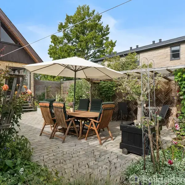 En terrasse med seks stole omkring et bord under en parasol. Stenbelægning danner gulvet, omgivet af grønne planter og træer, med et træhus og en murstensbygning i baggrunden.