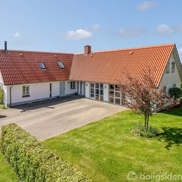 En hvid murstenshus med rødt tag står på en velplejet grund med frodige grønne arealer og en flisebelagt indkørsel. Et blomstrende træ og hække omkranser huset.