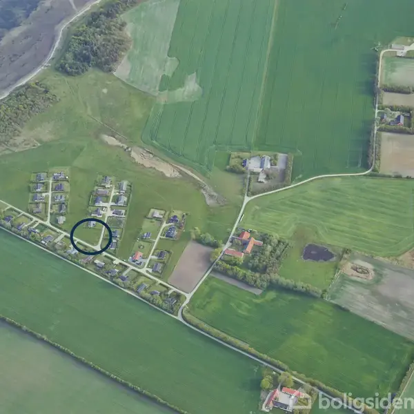 Et område med huse ses fra luften, omgivet af marker og en kystlinje. Et hus er markeret med en sort cirkel, og det ligger i et grønt landbrugsområde.