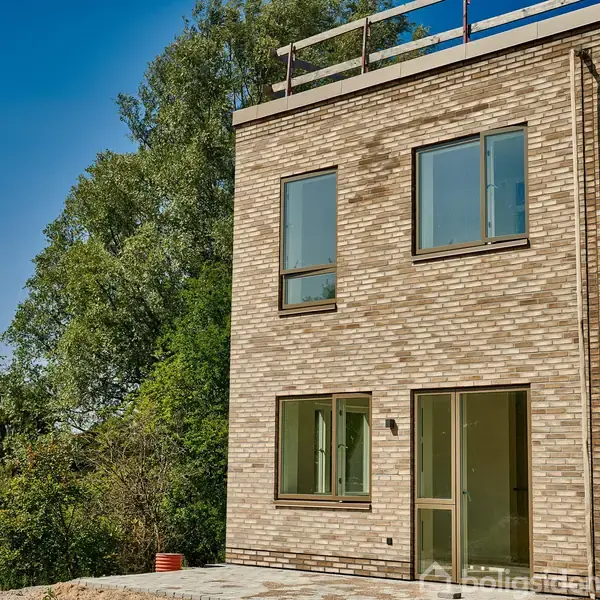 Et to-etagers hus med lyse mursten står ved siden af frodig vegetation. Vinduerne er store og moderne, og der er en terrasse udenfor. himlen er klar og blå.