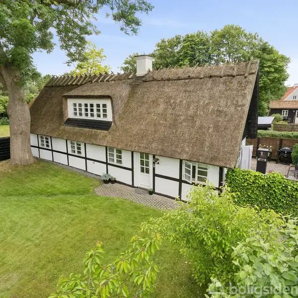 Et bindingsværkshus med stråtag står blandt træer og en velplejet græsplæne. Huset har hvide vægge med sorte bjælker og ligger i en grøn, landlig have.