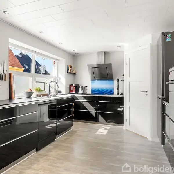 Et moderne køkken med sorte højglansskabe står lyst oplyst af et stort vindue. En komfur med indbygget ovn og en rustfri emhætte er placeret mellem skabene.