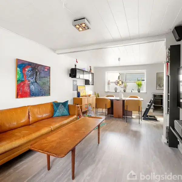 Brun lædersofa står i en stue med lyst trægulv, flankeret af et moderne vægmaleri. Foran står et træbord. Fjernsynet er monteret på en hvid væg, og vinduerne giver naturligt lys.