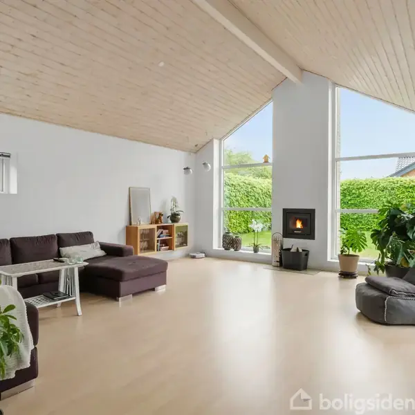 En stue med brun sofa står langs væggen. En brændeovn brænder ved et stort vindue, omgivet af grønne planter mod en havebaggrund. Lyse trægulve og skråt træloft.