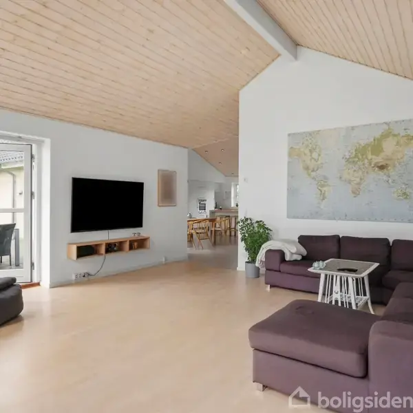 Stor stue med lyse trægulve og skråt loft. Brun hjørnesofa omkring et hvidt bord. Fladskærms-tv på væggen ved siden af. Udgang til terrasse gennem glasdør, verdenskort på væggen.