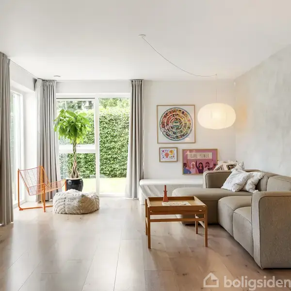 Stue med beige sofa, sofabord og væghængt TV. En plante står ved vinduet, som lader meget lys ind. Væggene er udsmykket med moderne kunstplakater.