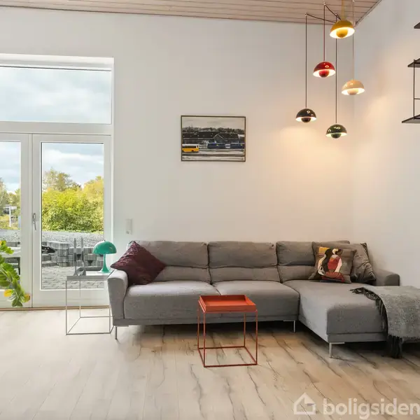 En grå sofa står i en moderne stue med træloft og lys træparket. En rød bordplade, hængende lamper, planter og en stor glasdør leder ud til en terrasse med grøn udsigt.