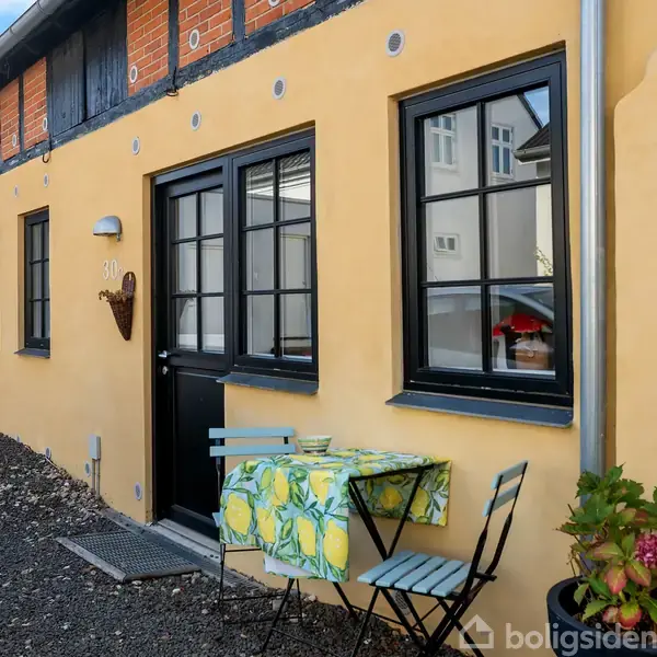 Et gult hus med sort dør og vinduer har et lille udendørs bord med citrondug og to stole foran. En grøn plante står til højre for døren. Nummeret "21" er synligt ved døren.