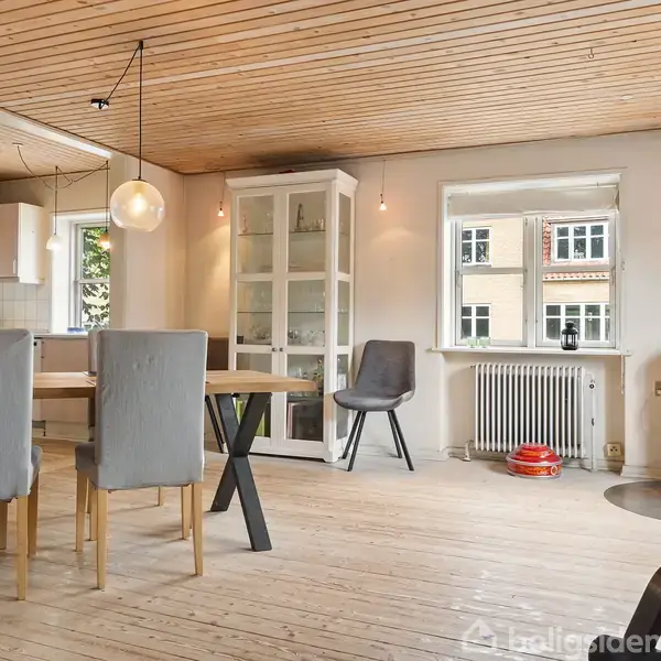 Spisebord med fire stole i en lys, træbeklædt stue. Vitrineskab står ved siden af vinduet. Brændeovn i hjørnet ved siden af en grå stol. Ingen tekst fundet.