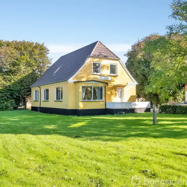 Gul villa står stille på en grøn græsplæne, omgivet af buske og træer. Huset har et mørkt tag og flere vinduer, der giver en lys og indbydende atmosfære.