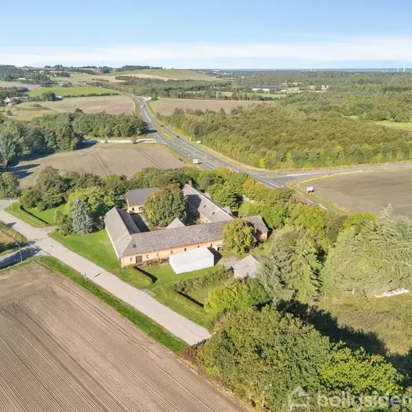 En gård med store bygninger ligger omgivet af grønne marker og træer. Veje snor sig gennem det åbne landskab. Området er landligt med skove og åbne marker i baggrunden.