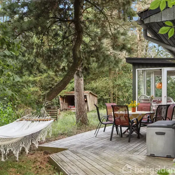 Hængekøje hænger fredeligt mellem træerne ved siden af en træterrasse. På terrassen er et bord med stole, omgivet af grønne buske og træer, i en skovbevoksning.