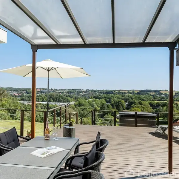 En terrasse med spisebord og sorte kurvestole under en halvtag. En åben parasol skaber skygge. Udsigt over en grøn dal med træer og huse i baggrunden.