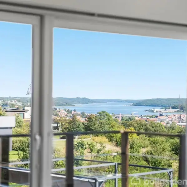 Udsigt gennem en glasdør til en terrasse med trægelænder. I baggrunden ses en by, grønne områder og en bugt under en klar blå himmel.